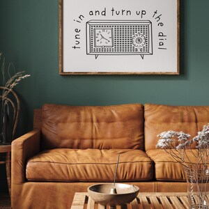 May include: A framed print with a black and white illustration of a vintage radio with the text "tune in and turn up the dial". The print is hanging on a dark green wall above a brown leather sofa.
