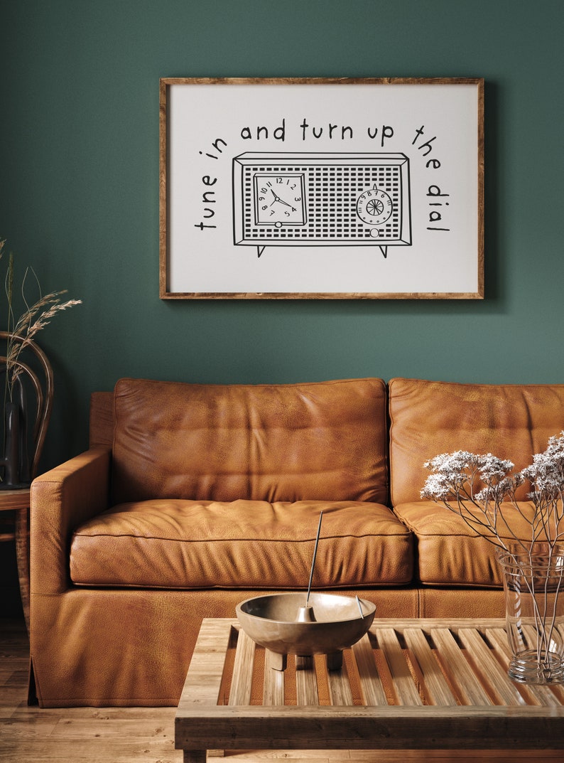 May include: A framed print with a black and white illustration of a vintage radio with the text "tune in and turn up the dial". The print is hanging on a dark green wall above a brown leather sofa.