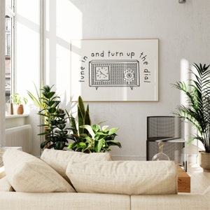 May include: A black and white illustration of a vintage radio with the text "tune in and turn up the dial". The illustration is framed and hanging on a white wall in a living room setting.