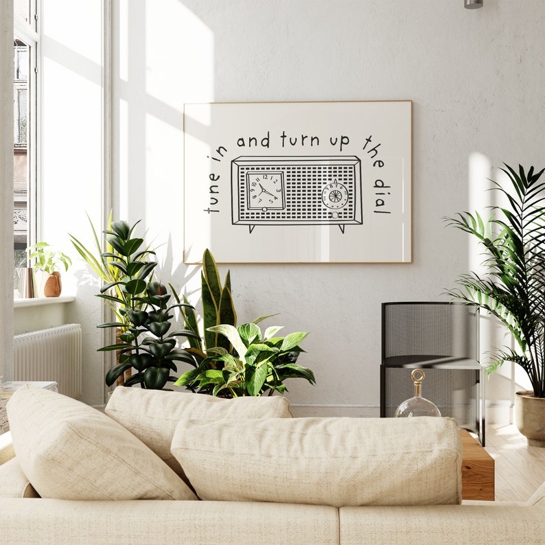 May include: A black and white illustration of a vintage radio with the text "tune in and turn up the dial". The illustration is framed and hanging on a white wall in a living room setting.