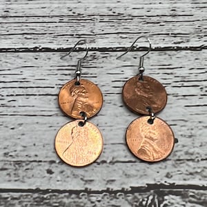 May include: Copper penny earrings with silver-colored ear wires. Each earring features two pennies, one from 2010 and one from 2014, and the other from 2012 and 2010. The earrings are displayed on a wooden surface.