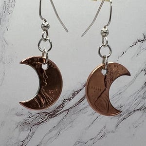May include: Copper-colored crescent moon earrings. Each earring is crafted from a penny, with the year 2017 visible on one. The earrings hang from silver-colored ear wires and small beads.