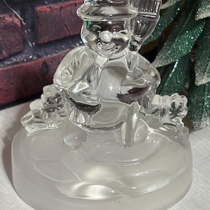 May include: A clear glass snowman figurine with a hat and broom. The snowman is sitting on a frosted, circular base. The background includes a brick wall and a green, glittery Christmas tree.