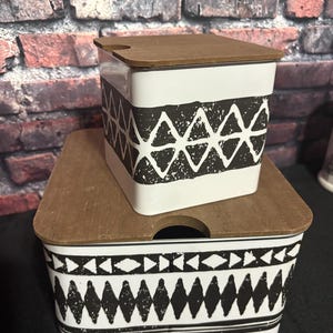 Puede incluir: Dos recipientes de cerámica blanca con tapas de madera. Los recipientes tienen patrones geométricos negros. Uno es cuadrado y el otro rectangular. Las tapas tienen un pequeño recorte. Los recipientes están apilados.