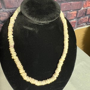 Può includere: Una collana color crema composta da piccole perline rotonde. La collana è esposta su un busto di velluto nero, che ne mette in risalto la forma e il design. Le perline sembrano uniformi per dimensioni e colore, creando un accessorio semplice ma elegante.
