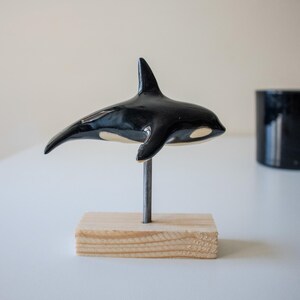 May include: A glossy black and white ceramic orca figurine mounted on a thin metal rod attached to a rectangular light-colored wooden base. The orca is positioned horizontally, showcasing its dorsal fin and characteristic white eye patch.