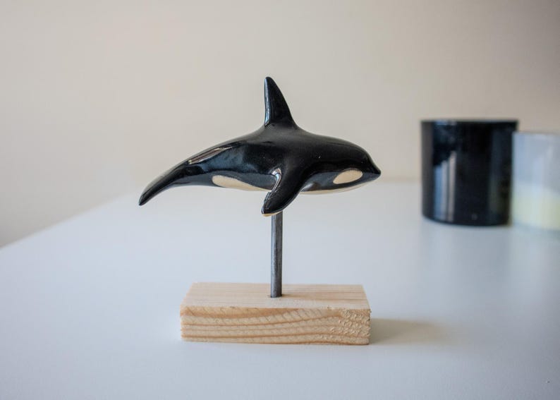 May include: A glossy black and white ceramic orca figurine mounted on a thin metal rod attached to a rectangular light-colored wooden base. The orca is positioned horizontally, showcasing its dorsal fin and characteristic white eye patch.