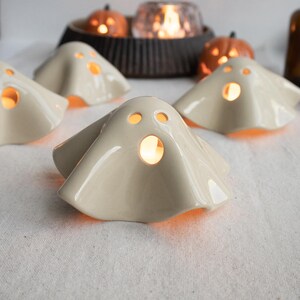 May include: A collection of off-white ceramic ghost-shaped tea light holders with cut-out eyes and mouths. Each ghost features a smooth, glossy finish and emits a warm orange glow from within, creating a festive atmosphere for Halloween decor.
