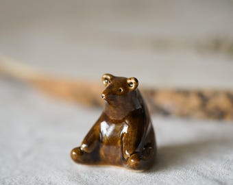 May include: A small, glossy, dark brown ceramic figurine of a seated bear. The bear has a smooth, reflective surface, rounded ears, and a pointed snout with small, dark, circular eyes. The figurine is sculpted in a sitting position.