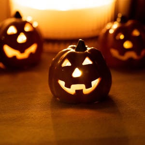Peut inclure: Trois citrouilles d'Halloween en céramique orange avec des yeux et des nez triangulaires sculptés ainsi que des bouches dentelées. Chaque citrouille possède une tige sombre au sommet et est éclairée de l'intérieur par une lumière chaude.