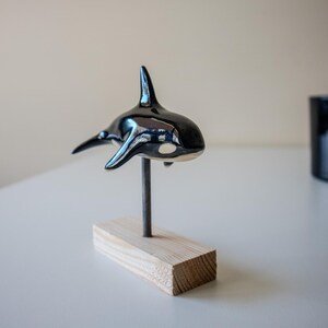 May include: A glossy black and white ceramic orca figurine mounted on a thin metal rod attached to a rectangular light-colored wooden base. The orca features a prominent dorsal fin, side pectoral fins, and a white patch near the eye area.