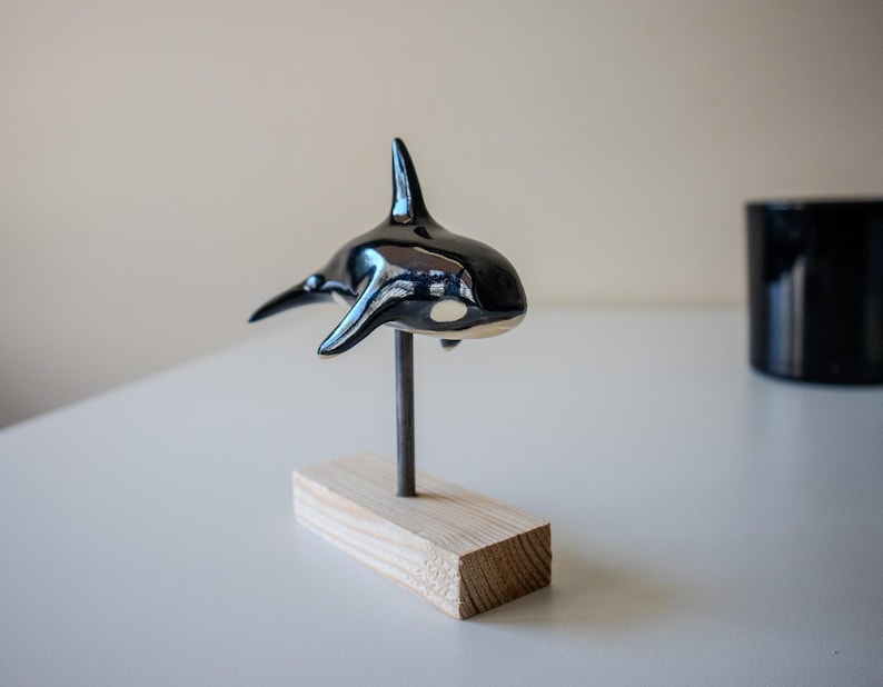 May include: A glossy black and white ceramic orca figurine mounted on a thin metal rod attached to a rectangular light-colored wooden base. The orca features a prominent dorsal fin, side pectoral fins, and a white patch near the eye area.
