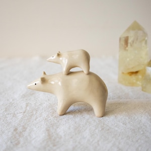 Könnte beinhalten: Eine glatte, cremefarbene Keramikskulptur, die einen größeren Bären zeigt, auf dessen Rücken ein kleinerer Bär steht. Beide Bären haben einfache, minimalistische Merkmale mit kleinen dunklen Augen und abgerundeten Ohren, überzogen mit einer Glasur.