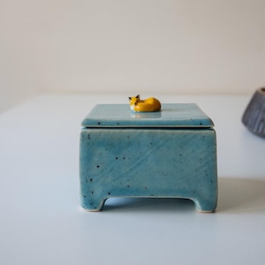 Może przedstawiać: Kwadratowe ceramiczne pudełko z jasnoniebieskim, nakrapianym szkliwem i czterema krótkimi nóżkami. Na pokrywce znajduje się mała, rzeźbiona figurka pomarańczowo-białego lisa. Pudełko ma gładkie, błyszczące wykończenie i prosty, funkcjonalny kształt.