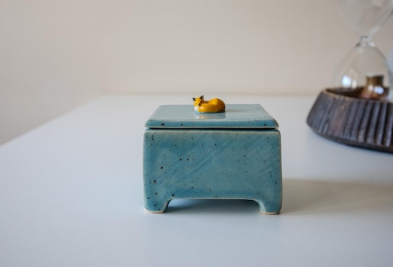 A hand-sculpted ceramic lidded box with a fox resting on top — the fox IS the lid, making it a tiny piece of woodland sculpture you can actually put rings in.