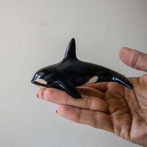 May include: A small, glossy ceramic figurine of an orca whale. The sculpture features the characteristic black and white coloration of an orca, with a smooth, reflective surface, a prominent dorsal fin, and white patches near the eyes and body.