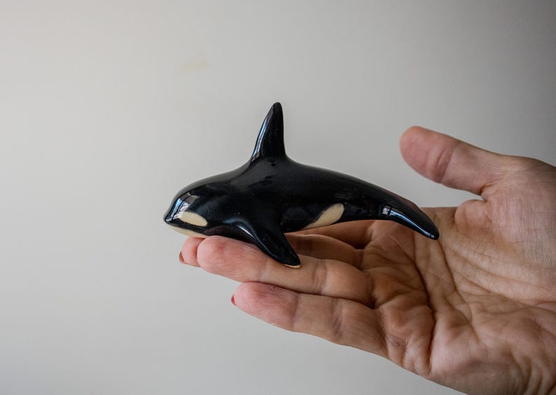 May include: A small, glossy ceramic figurine of an orca whale. The sculpture features the characteristic black and white coloration of an orca, with a smooth, reflective surface, a prominent dorsal fin, and white patches near the eyes and body.
