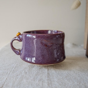 Puede incluir: Una taza de cerámica ancha de color morado con un acabado brillante y moteado, y una sola asa. Un pequeño detalle decorativo redondo de color naranja está fijado en la parte superior del asa. La taza tiene una forma redondeada y cónica.