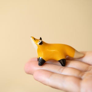 Puede incluir: Una pequeña figura de zorro de cerámica lisa con un cuerpo de color naranja vibrante, pecho y cara blancos, orejas con puntas negras y patas negras. El zorro está representado en posición de pie con la cabeza inclinada hacia arriba y acabado brillante.