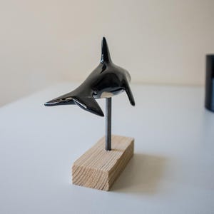 May include: A glossy black and white orca figurine mounted on a thin metal rod attached to a rectangular light-colored wooden base. The orca is positioned in a swimming pose, showcasing its dorsal fin, side fins, and tail fluke in a smooth finish.