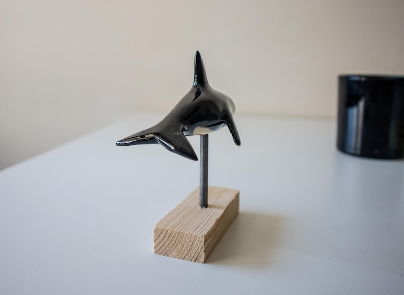 May include: A glossy black and white orca figurine mounted on a thin metal rod attached to a rectangular light-colored wooden base. The orca is positioned in a swimming pose, showcasing its dorsal fin, side fins, and tail fluke in a smooth finish.