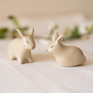 Może przedstawiać: Dwie małe, gładkie, beżowe ceramiczne figurki królików. Jeden królik stoi prosto ze spiczastymi uszami, podczas gdy drugi siedzi w pozycji spoczynkowej. Obie figurki mają proste, namalowane detale twarzy, w tym zamknięte oczy.