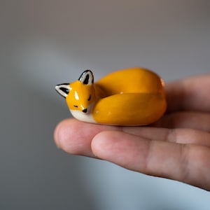 Könnte beinhalten: Eine kleine, glatte Keramikfigur eines zusammengerollten Fuchses. Der Fuchs ist überwiegend orange mit weißem Bauch und Gesicht, schwarzen Ohrspitzen und geschlossenen Augen. Die glänzende Oberfläche reflektiert das Licht und betont die Form.