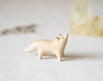 May include: A small, smooth, cream-colored ceramic fox figurine standing in a profile position. The fox has a glossy finish, pointed ears, and a slender tail, with its head tilted slightly upward and eyes depicted as simple, thin horizontal lines.