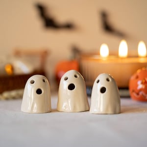 Peut inclure: Trois petites figurines de fantômes en céramique blanc cassé aux formes lisses et arrondies. Chaque fantôme présente deux découpes circulaires pour les yeux et une découpe circulaire plus grande pour la bouche, créant une expression ouverte.