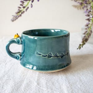 May include: A teal ceramic mug with a glossy finish, featuring a small yellow accent on the handle and a horizontal embossed branch design on the side. The mug has a wide, rounded body and a sturdy handle, showcasing a smooth, glazed surface.