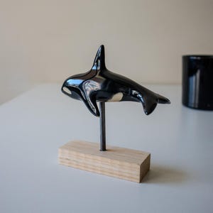 May include: A glossy black and white ceramic orca figurine mounted on a thin metal rod attached to a rectangular light-colored wooden base. The orca is depicted in a swimming position with its dorsal fin upright and tail flukes extended horizontally.