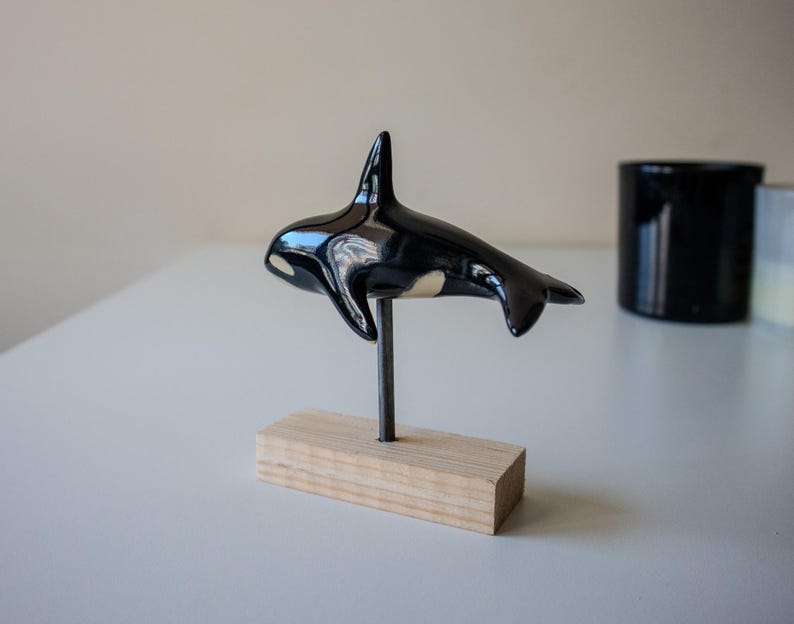 May include: A glossy black and white ceramic orca figurine mounted on a thin metal rod attached to a rectangular light-colored wooden base. The orca is depicted in a swimming position with its dorsal fin upright and tail flukes extended horizontally.