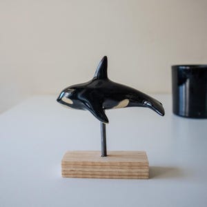 May include: A glossy black and white ceramic orca figurine mounted on a thin metal rod attached to a rectangular light-colored wooden base. The orca features a prominent dorsal fin, a white eye patch, and a smooth, reflective surface finish.