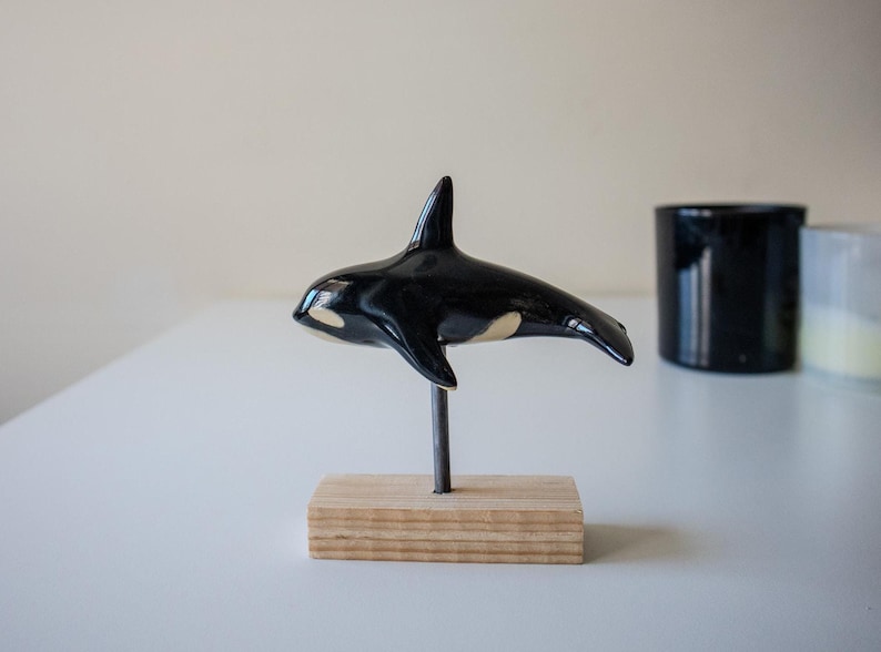 May include: A glossy black and white ceramic orca figurine mounted on a thin metal rod attached to a rectangular light-colored wooden base. The orca features a prominent dorsal fin, a white eye patch, and a smooth, reflective surface finish.