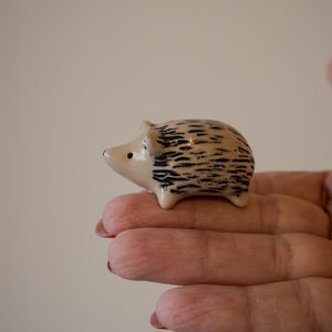 Peut inclure: Une petite figurine de hérisson en céramique beige avec une finition lisse et brillante. Le hérisson présente des marques noires en forme de tirets peintes à la main sur le dos pour représenter les piquants, un museau pointu et un œil noir.