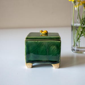Puede incluir: Una caja cuadrada de cerámica de color verde oscuro con un acabado brillante y cuatro patas cortas de color claro. La tapa presenta una pequeña figura de animal amarilla esculpida en la parte superior. La caja tiene una superficie lisa.
