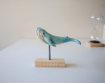 May include: A light blue ceramic whale sculpture with a smooth, glossy finish and visible dark vertical lines along its underside. The whale is mounted on a thin, dark metal rod attached to a rectangular, light-colored wooden base.