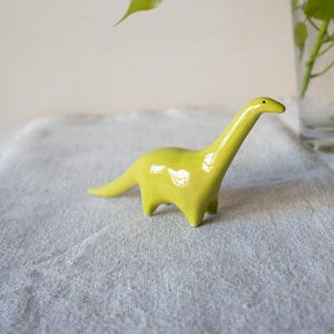 Könnte beinhalten: Eine kleine, glänzend limettengrüne Keramikfigur in Form eines Dinosauriers mit langem Hals. Die Figur hat eine glatte, reflektierende Oberfläche, einen schlanken Hals, einen spitzen Schwanz und vier kurze Beine sowie einen kleinen schwarzen Punkt als Auge.