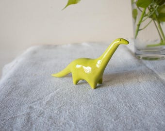 May include: A small, glossy, lime-green ceramic figurine shaped like a long-necked dinosaur. The figure features a smooth, reflective surface, a slender neck, a pointed tail, and four short legs, with a single small black dot representing the eye.