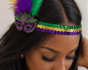 May include: A headband featuring three rows of sequins in green, gold, and purple. Attached to the side is a decorative mask applique adorned with small beads in matching colors, accented by a cluster of upright purple, green, and yellow feathers.
