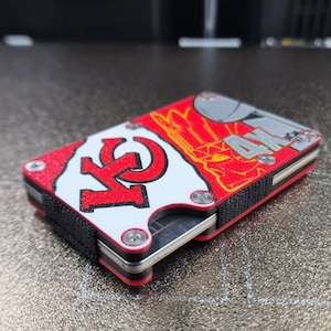 May include: A red, white, and black wallet featuring the Kansas City Chiefs logo and a football-themed design. The wallet is made of a durable material and has a black elastic band to hold the contents securely.