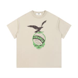 May include: Cream-colored t-shirt featuring a graphic of a black eagle perched atop a green and white skull design. The skull has the word "BRANCA" in a circular font. The shirt has a crew neck and short sleeves.