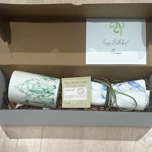 May include: A gift box containing a white mug with green floral design, a folded blue and white patterned cloth, a tea packet, and a greeting card that reads "Happy Birthday! Thanna". The box is lined with brown paper.