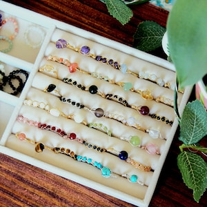 May include: A white tray with rows of gemstone rings. Each ring is made with a different type of gemstone, including amethyst, rose quartz, turquoise, and lapis lazuli. The rings are displayed on a white background.