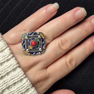 May include: A silver ring with a square, decorative design. The ring has a central red stone, surrounded by blue, yellow, and green enamel, and silver detailing. The ring is on a hand, with a cream-coloured sweater sleeve visible.