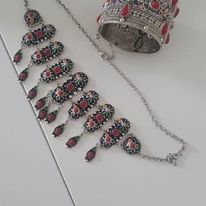 Puede incluir: Juego de joyas plateadas con collar y pulsera de puño. El collar tiene una cadena con colgantes ovalados decorativos en azul, rojo, verde y amarillo. La pulsera de puño tiene detalles de piedras rojas.