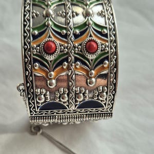 Puede incluir: Una pulsera de puño plateada con intrincados patrones florales y geométricos. La pulsera presenta detalles de esmalte rojo, verde, azul y naranja, y está adornada con cabujones de piedras preciosas rojas. El cierre es visible con un pasador plateado.