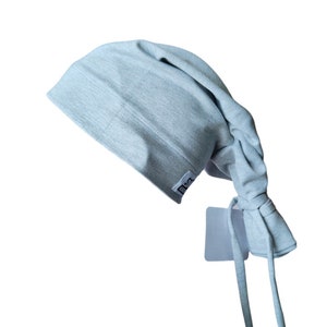 Puede incluir: Un gorro de quirófano de algodón gris claro con una pequeña etiqueta blanca que dice "Elsa". El gorro tiene lazos largos que se pueden atar en la parte posterior del cuello.