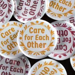 May include: A collection of white round buttons with the text "Care for Each Other" in orange. Each button is encircled by a decorative orange floral wreath. Some buttons have the same text in a burgundy color.
