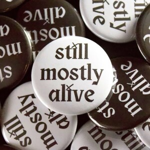 May include: A collection of round buttons with the phrase "still mostly alive" printed in black on a white button. Other buttons are black with the same phrase in white. The buttons are scattered.